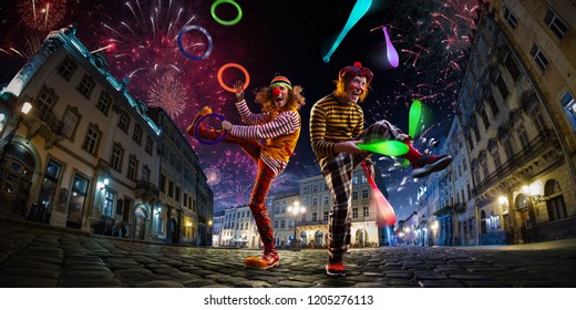 Night Street Circus Performance Whit Two Clowns, Juggler. Festival City Background. Fireworks And Celebration Atmosphere.