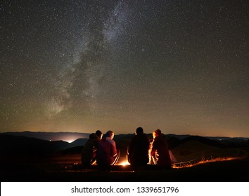Night summer camping in the mountains. Back view group of four friends tourists sitting on a bench made of logs together around campfire under amazing night starry sky full of stars and Milky way.