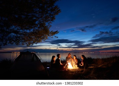 Night summer camping on lake shore. Group of five young happy tourists sitting in high grass around bonfire near tent under beautiful blue evening sky. Tourism, friendship and beauty of nature concept - Powered by Shutterstock