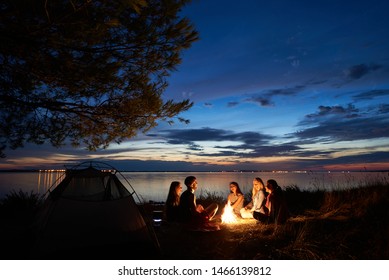 Night summer camping on sea shore. Group of young tourists sitting, laughing in high grass around bonfire near tent under beautiful blue evening sky. Tourism, friendship and beauty of nature concept. - Powered by Shutterstock