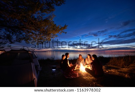 Night summer camping on sea shore. Group of five young tourists sitting on the beach around campfire near tent under beautiful blue evening sky. Tourism, friendship and beauty of nature concept.