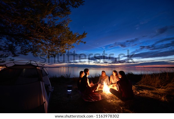 Night summer camping on sea shore. Group of five\
young tourists sitting on the beach around campfire near tent under\
beautiful blue evening sky. Tourism, friendship and beauty of\
nature concept.