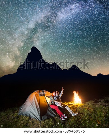 Night tent camping. Charming pair sitting near tent and campfire and enjoying incredibly beautiful starry sky in the background silhouette of the mountains. Long exposure