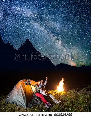 Night tent camping. Young pair sitting near tent and campfire and enjoying incredibly beautiful starry sky in the background silhouette of the mountains. Long exposure