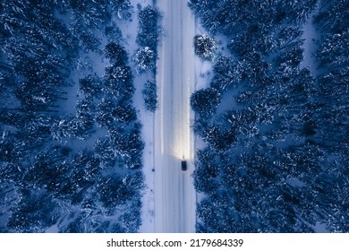 Night time aerial view of snowy road in pine tree forest in winter season. Drone top down view of snowy winter road surrounded pine and fir forest and car with light
