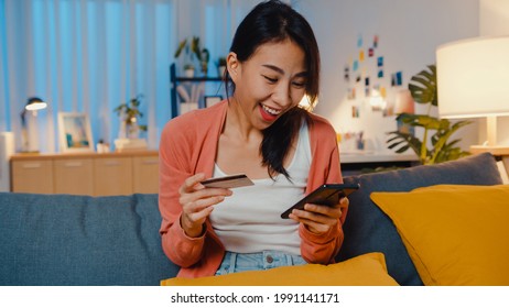 Night Time Beautiful Smiling Asian Lady Use Cellphone Order Online Shopping Product With Credit Card On Sofa In Living Room. Stay At Home, Self Quarantine Activity, Fun Activity For Covid Quarantine.