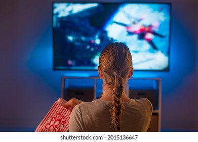 Night Time A Young Woman With Long Blond Hair Is Watching A Movie On TV With Interest. She Is Holding A Box Of Popcorn. Rest, Entertainment. There Is A Blue Neon Light In The Room.