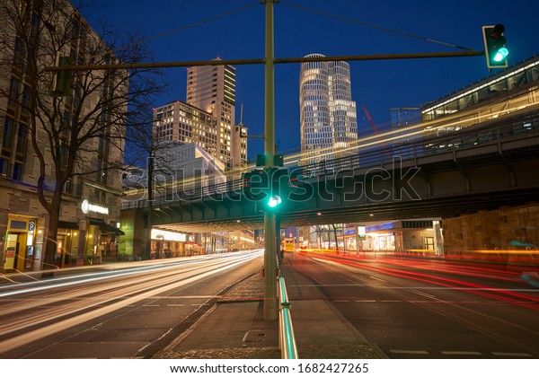Night traffic on the german city street. Red and\
white light trails created due to long exposure. skyscraper, office\
buildings and a bridge in background, traffic light in the front,\
blue sky, dusk