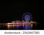 Night view of a Ferris wheel illuminated with vibrant lights reflecting on the water, located at a waterfront entertainment complex.