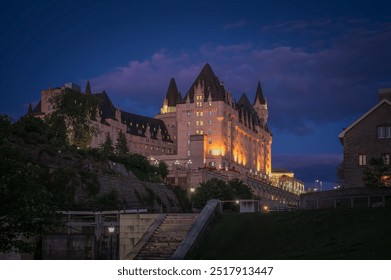 Night view of the iconic Fairmont Château Laurier hotel illuminated under twilight blue hour sky, dramatic purple clouds, above the historic Rideau Canal locks, Ottawa, Ontario, Canada (August 2022)