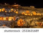 Night view of illuminated ancient cave dwellings carved into a rocky hillside, with modern buildings visible above.