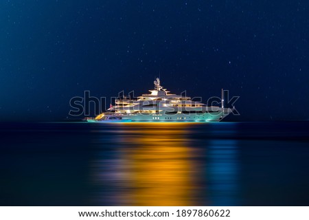 Night view to large illuminated white boat located over horizon, colorful lights coming from yacht reflect on the surface of the the Gulf sea. Shot at blue hour.  