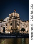 Night view of the Reichstag building in Berlin, Germany, with a crowd gathered by the river and the German flag illuminated.