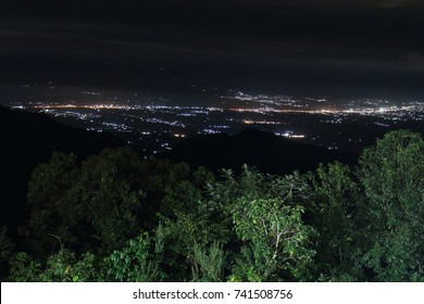 Night View From A Top Of Mountain, Light Of The City At The Below Is Bright In The Dark. Green Forest In Foreground