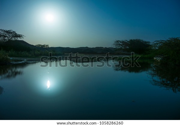 Night waterscape on\
lake coast with sky moon and forest reflection on water surface.\
Low light scenery.