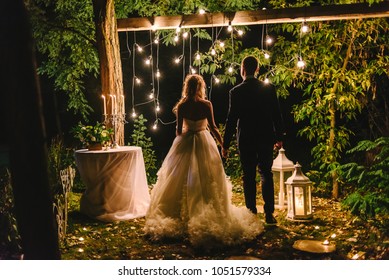 Night wedding ceremony with candles, lanterns and lamps on tree. Bride and groom holding hands on background of bulb lights, back view. Beautiful young couple standing under tree at night, copy space