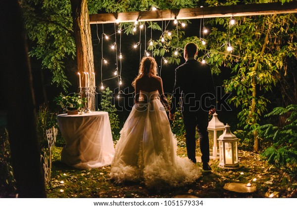 Night wedding ceremony with candles, lanterns and\
lamps on tree. Bride and groom holding hands on background of bulb\
lights, back view. Beautiful young couple standing under tree at\
night, copy space