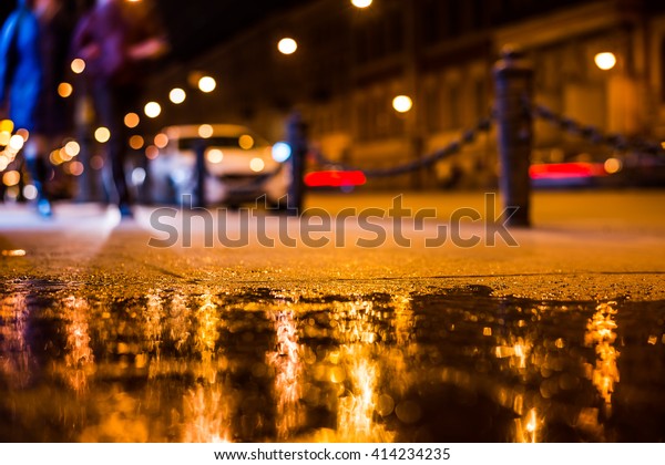 Nights lights of the big city, a\
couple of people walking along the main street of the city. Close\
up view from the level of the sidewalk, focus on the\
sidewalk