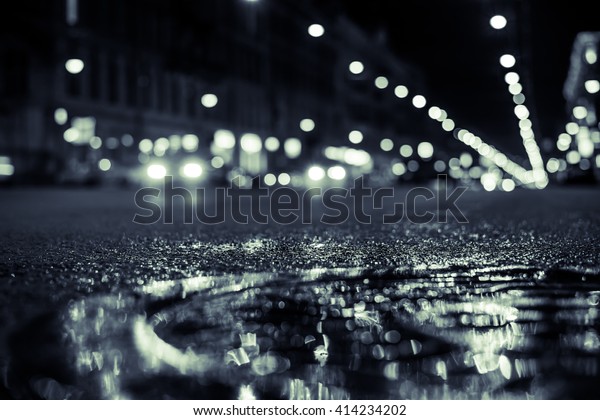 Nights lights of the big\
city, the main city street in rushhour. Close up view of a puddle\
on the level of the hatch, image in the green-blue toning, focus on\
the asphalt