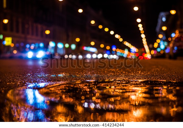 Nights lights of the big city, the main city\
street in rushhour. Close up view of a puddle on the level of the\
hatch, focus on the\
asphalt