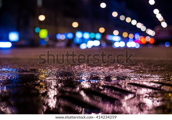 Nights lights of the big city, the main city\
street in rushhour. Close up view of a puddle on the level of the\
hatch, in blue tones, focus on the\
asphalt