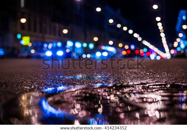 Nights lights of the big city, the main city\
street in rushhour. Close up view of a puddle on the level of the\
hatch, in blue tones, focus on the\
asphalt