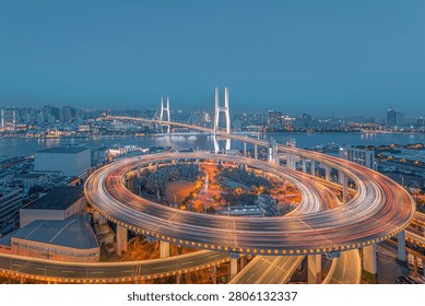 Nighttime long-exposure view of a modern cable-stayed bridge and spiral elevated highway interchange in Shanghai, with illuminated city skyline and river in background.