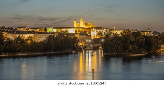 A nighttime view of Prague Castle illuminated across the Vltava River. The castle spires and buildings are reflected in the water, creating a picturesque scene. The cityscape is bathed in warm, golden - Powered by Shutterstock