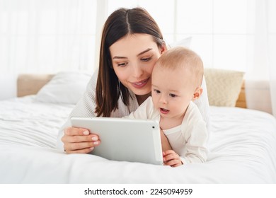 Nine-month-old baby child with mom are sitting on the bed and looking at the screen of digital tablet. Child development using modern technology. The child is talking on a video call. Online learning - Powered by Shutterstock