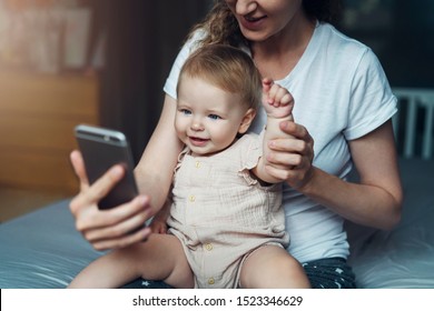 Nine-month-old Baby Girl Sits With Her Mother On Bed And Looks On Smartphone Cartoons, Plays Game. Child Is Talking To Her Grandmother Via Video Link. Mom Shows Her Daughter Pictures On Phone Screen.