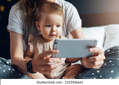 Nine-month-old baby girl sits with her mother on bed and looks on smartphone cartoons, plays game. Child is talking to her grandmother via video link. Mom shows her daughter pictures on phone screen.