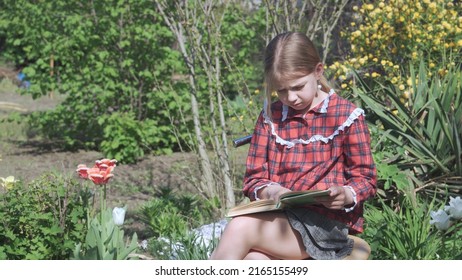 A Nine-year-old Girl In A School Uniform Sits In The Garden And Carefully Reads A Book. High Quality Photo