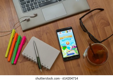 Nitra, Slovakia, April 3, 2017: Pacman Bounce Application In A Mobile Phone Screen. Workplace With A Laptop, An Earphones, Notepad, Pen, Tea, Sunglasses And Color Markers On Wooden Background