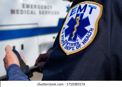 NJ / USA - April 12, 2020: EMT patch on man's arm sleeve while man plays on phone with white ambulance in background during 2020 COVID-19 Pandemic