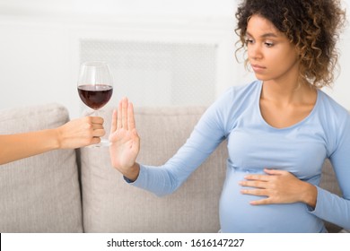 No alcohol. Pregnant woman gesturing stop to offered glass of wine, sitting on sofa