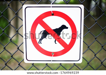 No dog allowed square sign with red circle and black dog attached to a chain link fence in a garden zone or park with trees in the background