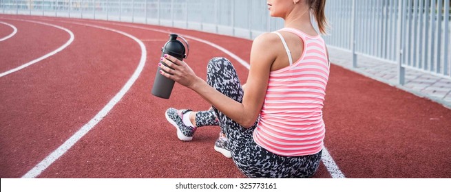 No face. Cute slim young adult girl hold in hand black plastic shaker. Blonde woman sitting on running track and drinking bottled water. Empty sporty space for inscription or other objects and people - Powered by Shutterstock