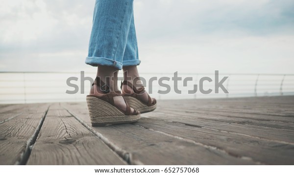 No face. Female feet are walking along the wooden\
embankment near the sea or ocean. Girl in stylish woven sandals on\
a platform and jeans.