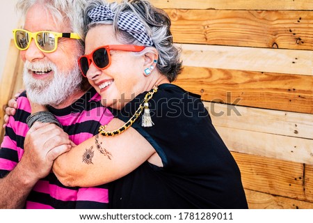 No limit age with nice and funny elderly caucasian couple have fun and smile in a portrait with wooden background - trendy and coloured accessories on old cheerful and happy people