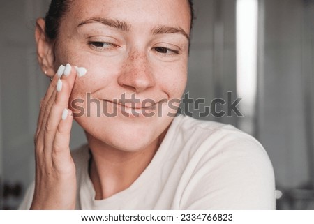 No make up woman applying cream on her face with a smile, home bathroom
