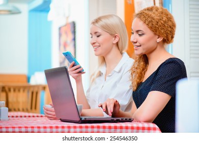 No Minute Without Laptop. Side View Of Two Young Women Working On Laptop And Looking At Mobile Phone While Sitting On The Couch Of The Cafe