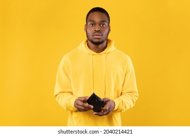 No Money. Shocked Black Guy Showing His Empty Wallet Looking At Camera Standing Over Yellow Background. Poor African American Millennial Man Having Difficulties Struggling During Financial Crisis