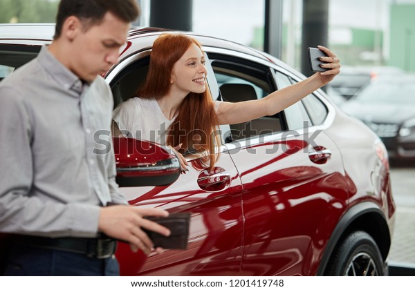 No money. Young man near new car\
looking empty pocket, while his wife doing selfie in\
car