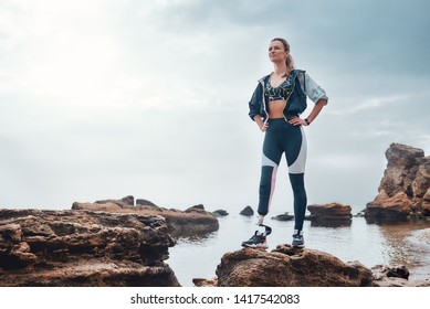 No pain, no gain. Confident disabled woman in sportswear with bionic leg standing on a stone in front of the sea and thinking about life goals. - Powered by Shutterstock