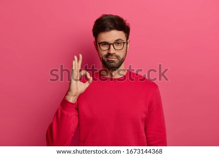 No problem concept. Bearded man makes okay gesture, has everything under control, all fine gesture, wears spectacles and jumper, poses against pink background, says I got this, guarantees something