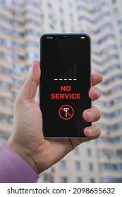 'No Service' Notification On The Cell Phone Screen. Smartphone In Man's Hand Against The Background Of A Residential Skyscraper. The Cellular Signal Is Lost. Denial Of Service. Technological Problem