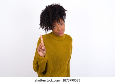 No sign gesture. Closeup portrait unhappy Young beautiful African American woman wearing knitted sweater against white wall raising fore finger up saying no. Negative emotions facial expressions, feel