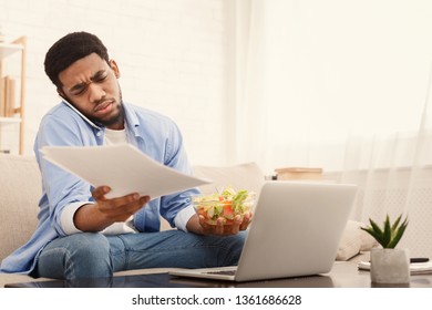 No Time For Break. Nervous African American Guy Eating Healthy Breakfast, Talking On Mobile Phone And Working On Laptop At Home, Copy Space