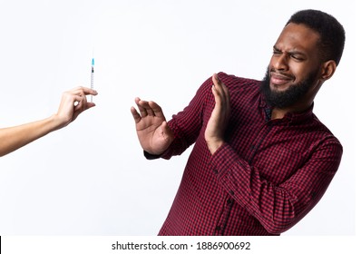 No Vaccination. Scared African Man Gesturing Stop To Hand Offering Syringe With Vaccine Refusing To Be Vaccinated Standing Over White Studio Background. Needle Fobia, Fear Of Covid-19 Vaccines