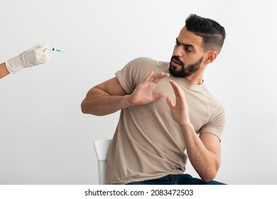 No Vaccination. Scared Young Arab Man Gesturing Stop To Doctor Offering Syringe With Covid Vaccine, Refusing To Be Vaccinated On White Studio Background. Needle Fobia, Fear Of Coronavirus Immunization